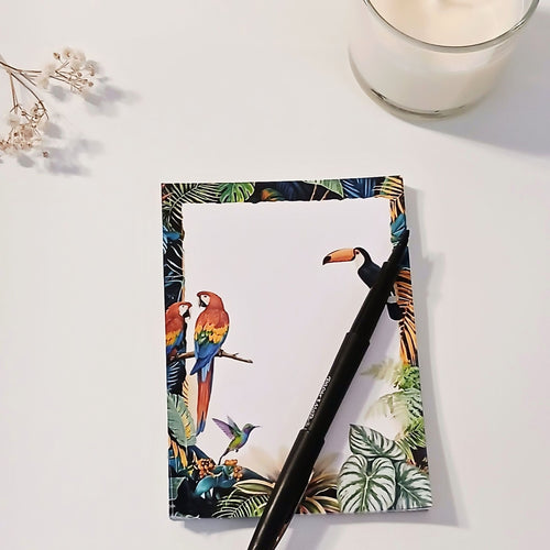 Bloc-notes A6 Maximalisme Tropical, main qui écrit au feutre sur la page blanche, perroquets rouges et toucan dans le cadre