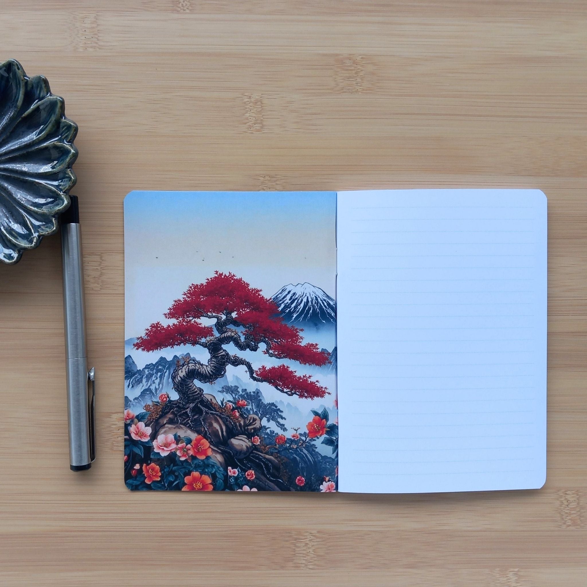 Ouverture d'un petit carnet d'écriture sur un plan de travail en bambou, révélant sa double page intérieure. La page de droite est lignée pour les notes. La page de gauche est une œuvre d'art à part entière, illustrant un bonsaï tortueux au feuillage vermillon, des fleurs rouges sauvages, une montagne enneigée et un vol d'oiseaux dans un ciel bleu pâle.