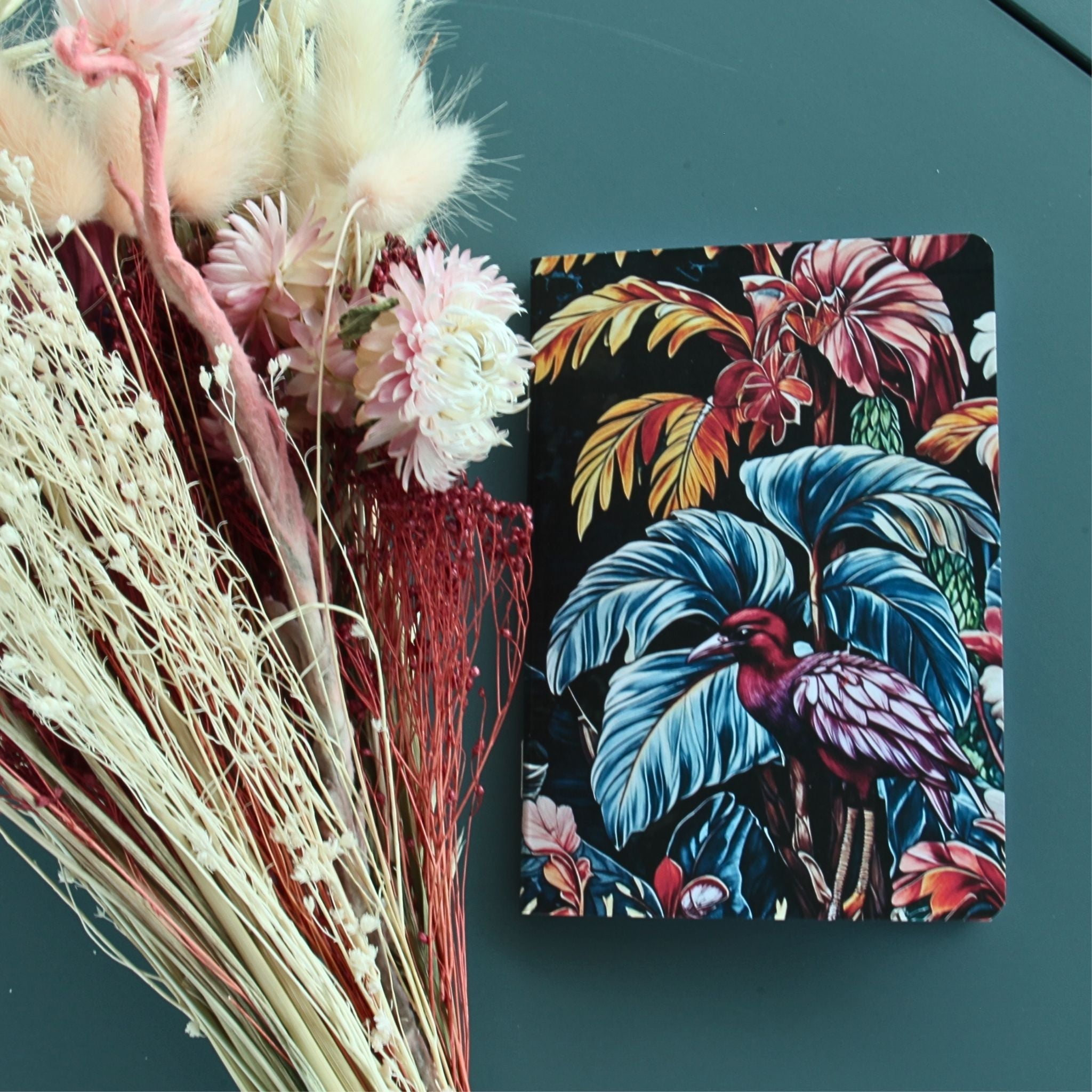 Gros plan sur le carnet à motif végétal luxuriant. Design floral sombre aux nuances émeraude et rose poudré, mettant en scène un oiseau exotique caché dans un feuillage tropical dense.