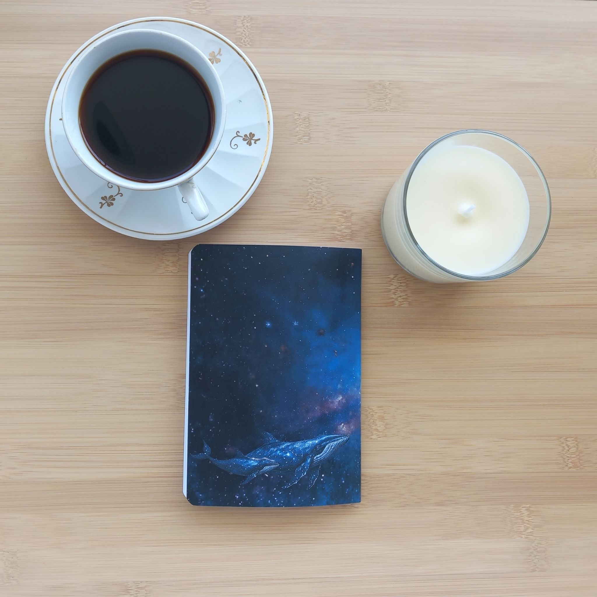 Vue de dessus du carnet fermé "L'Odyssée Stellaire" posé sur une table en bambou, entre une tasse de café et une bougie blanche. La quatrième de couverture illustre deux baleines (une grande et un baleineau) nageant dans l'espace profond d'une nébuleuse bleu nuit étoilée.