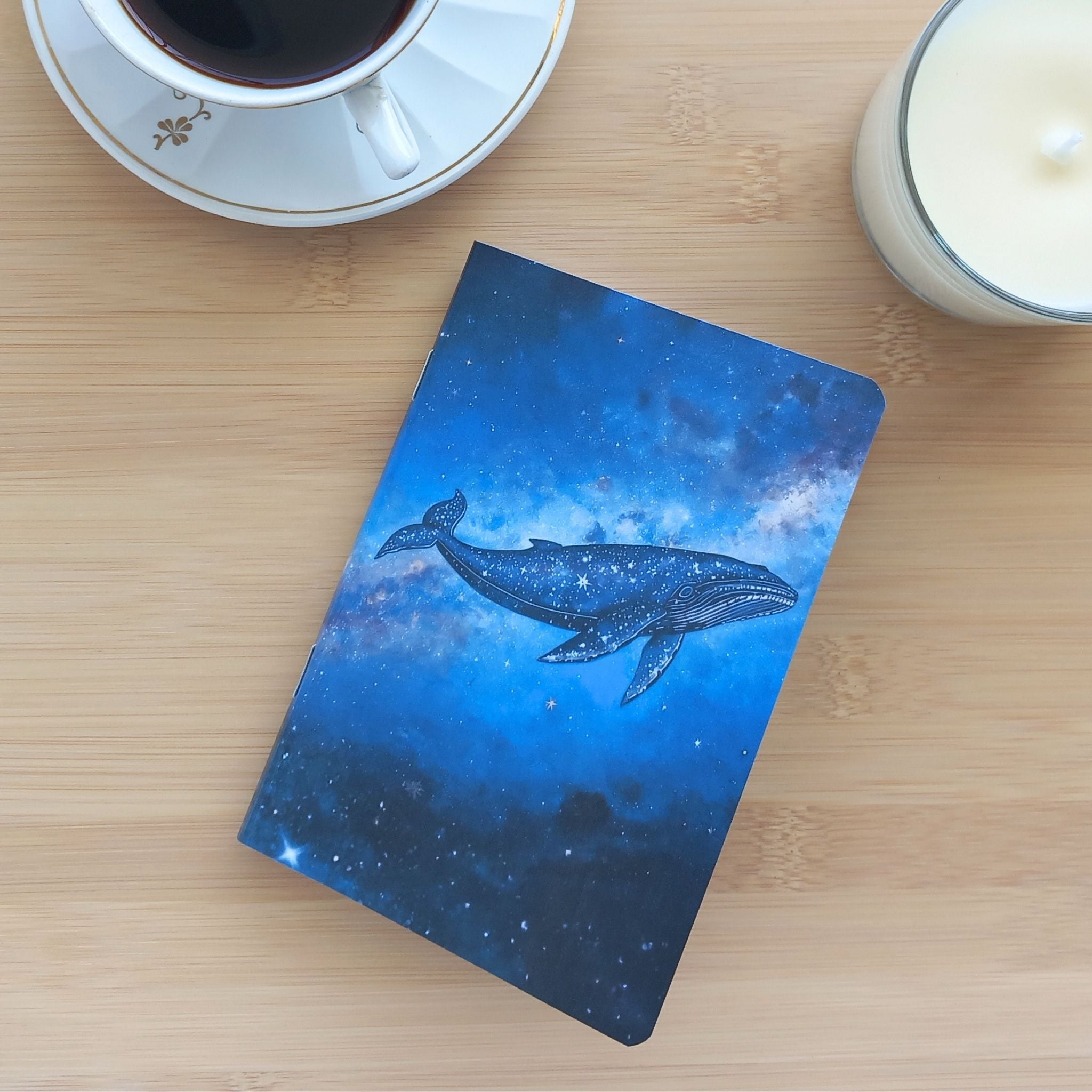Première de couverture du carnet de notes A6 fait main, photographié de biais sur un fond en bois clair avec un café et une bougie. L'illustration centrale met en valeur une grande baleine onirique traversant une galaxie lumineuse aux nuances bleu turquoise et outremer.