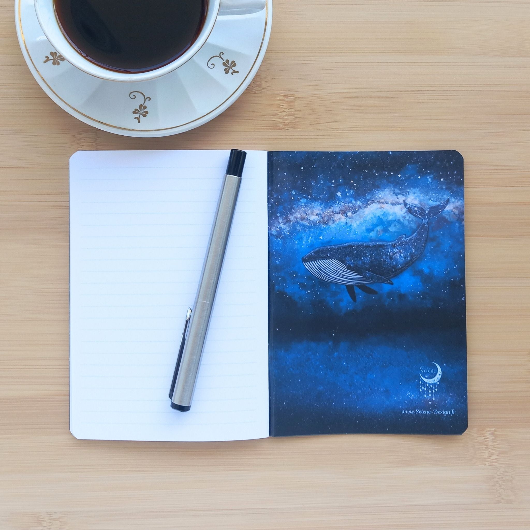 Carnet ouvert révélant sa page de garde arrière illustrée d'une baleine plongeant dans un ciel étoilé avec le logo céleste Sélène Design. À gauche, un stylo en métal argenté est posé sur les pages lignées vierges.