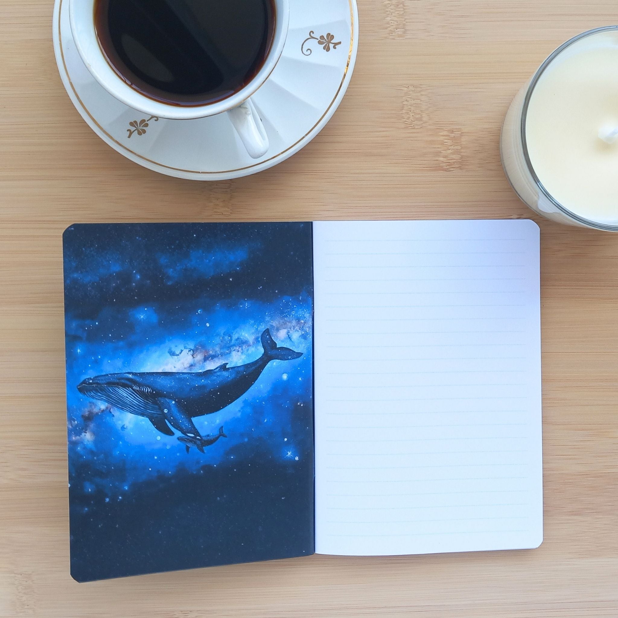 Carnet d'écriture ouvert posé sous une tasse de café et une bougie. La page de garde de gauche révèle une illustration pleine page d'une mère baleine et de son petit dans le cosmos. La page de droite montre le papier intérieur blanc ligné.