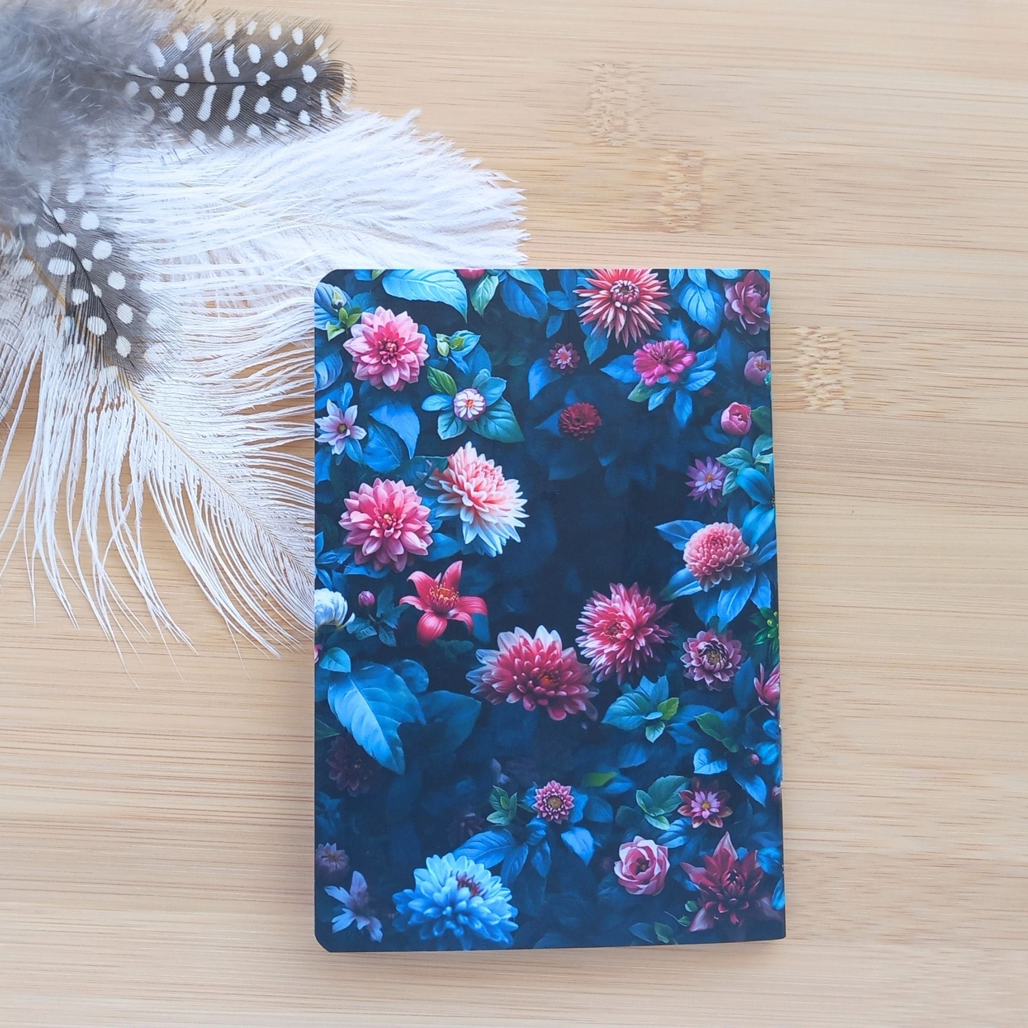 Vue de la quatrième de couverture du bloc-notes "L'oiseau et le Jardin Nocturne". Le motif au dos présente un dense feuillage bleuté parsemé de dahlias et de fleurs roses vibrantes, photographié près de plumes douces.
