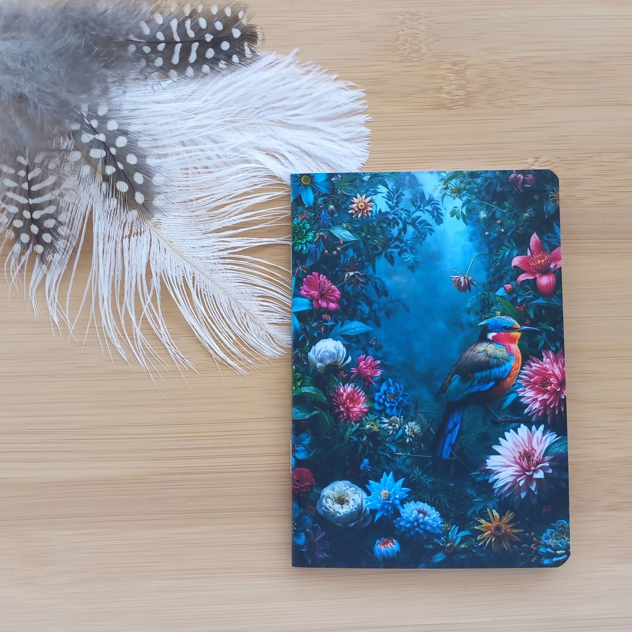 Première de couverture du petit carnet artisanal posé sur une surface en bambou à côté de plumes décoratives claires et tachetées. L'illustration centrale met en valeur un oiseau au plumage vif entouré d'un jardin floral nocturne luxuriant.