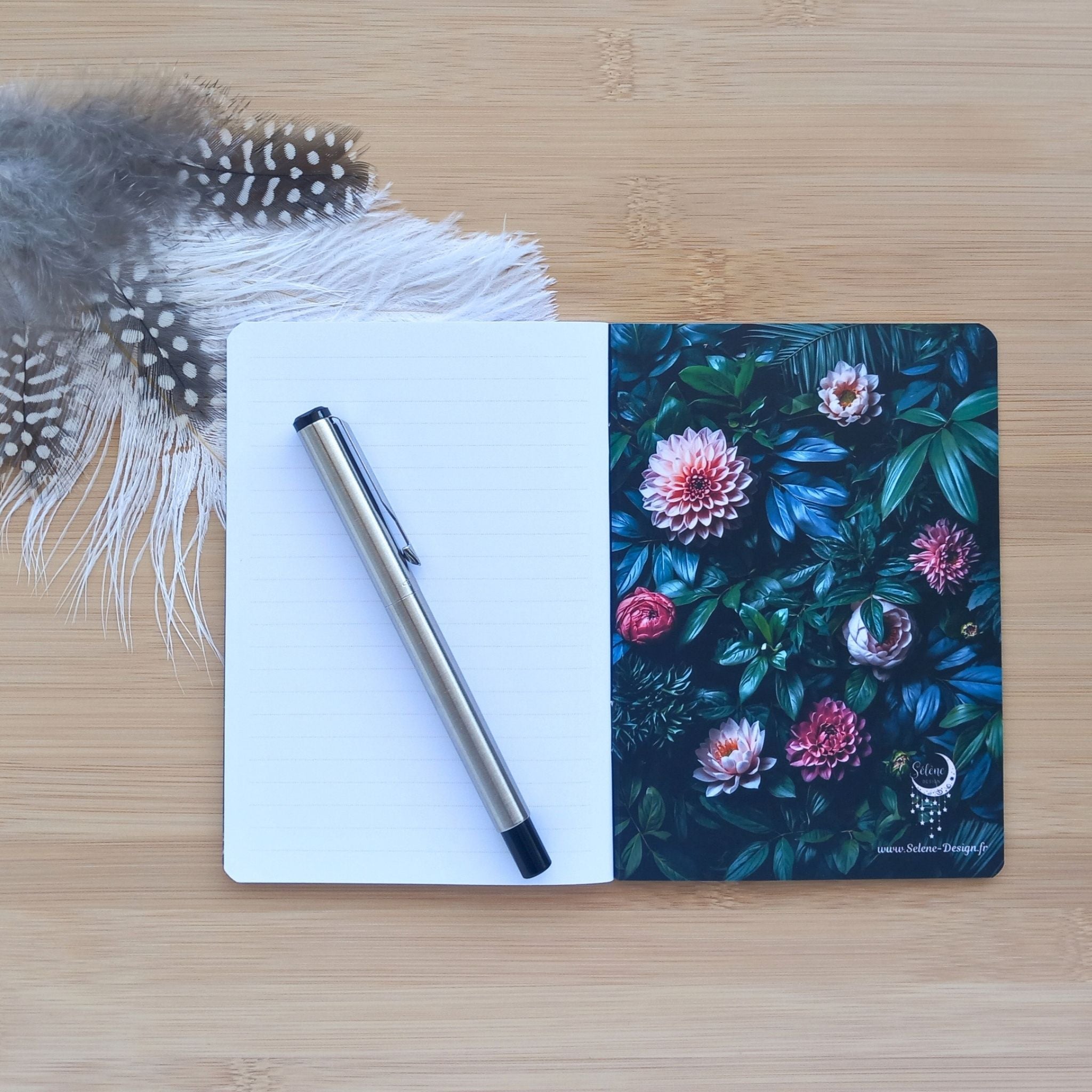 Carnet ouvert avec un stylo argenté reposant sur la page lignée vierge de gauche. La page de garde droite dévoile des feuillages sombres, de grandes fleurs roses et le logo lunaire Sélène Design.