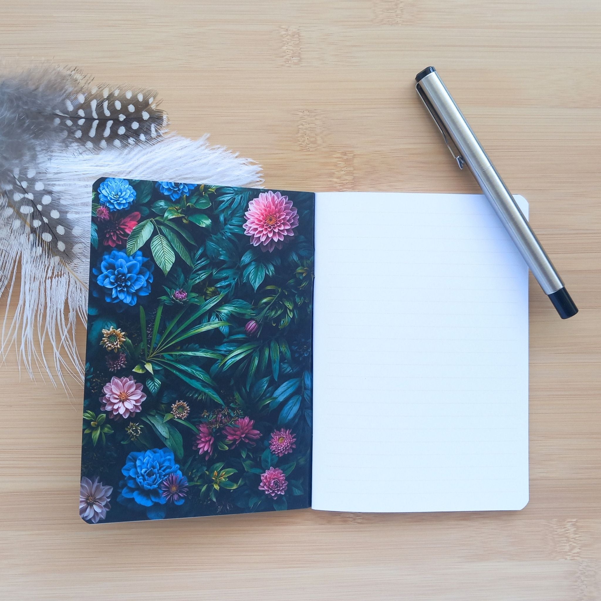 arnet d'écriture ouvert révélant la couverture intérieure gauche décorée d'un motif floral sombre avec des fleurs bleues et roses. Sur la page de droite, le papier blanc ligné est visible avec un stylo en métal posé en diagonale.