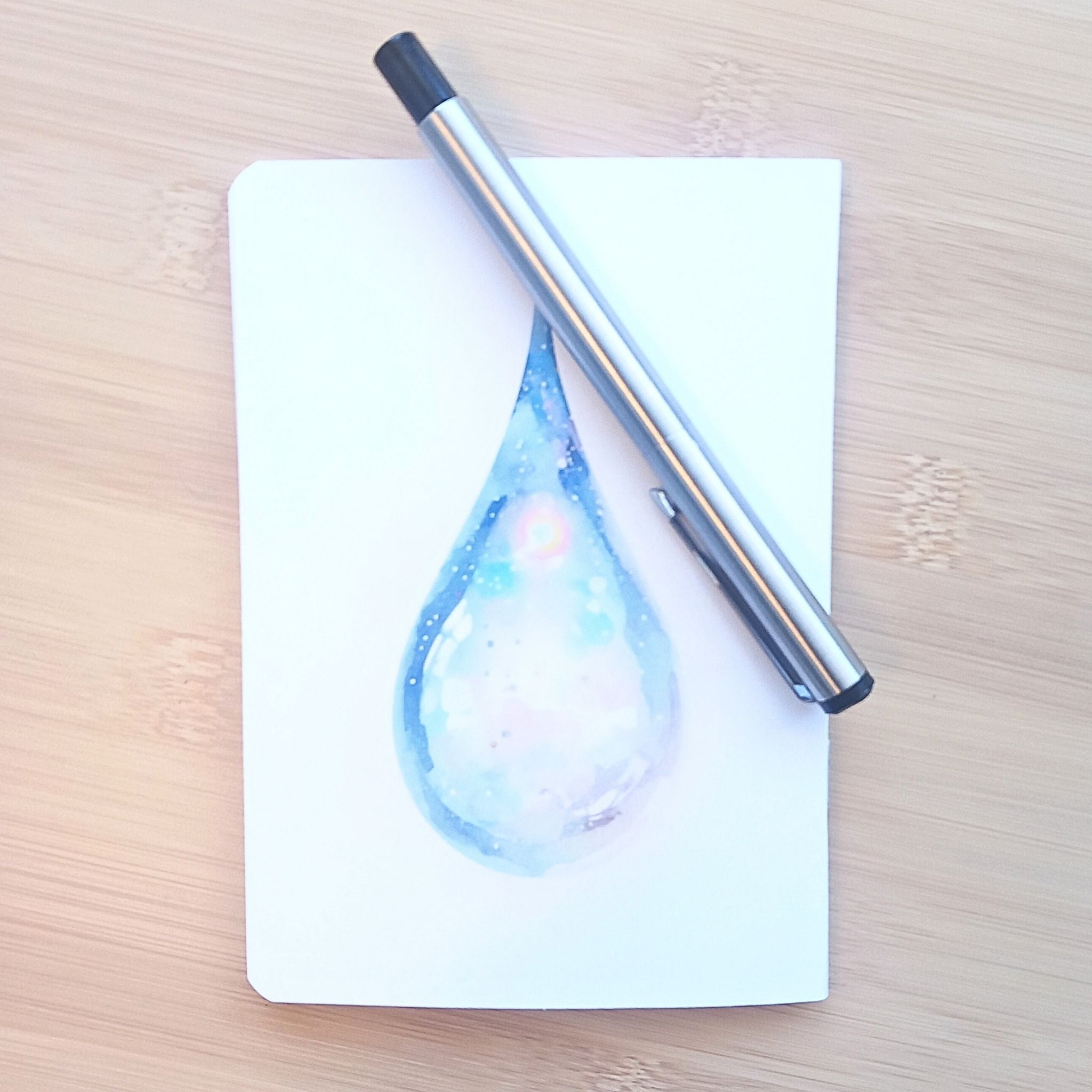Dos du carnet de notes A6 "Le colibri et la rosée", décoré d'une grande goutte d'eau irisée peinte à l'aquarelle dans des tons pastel.