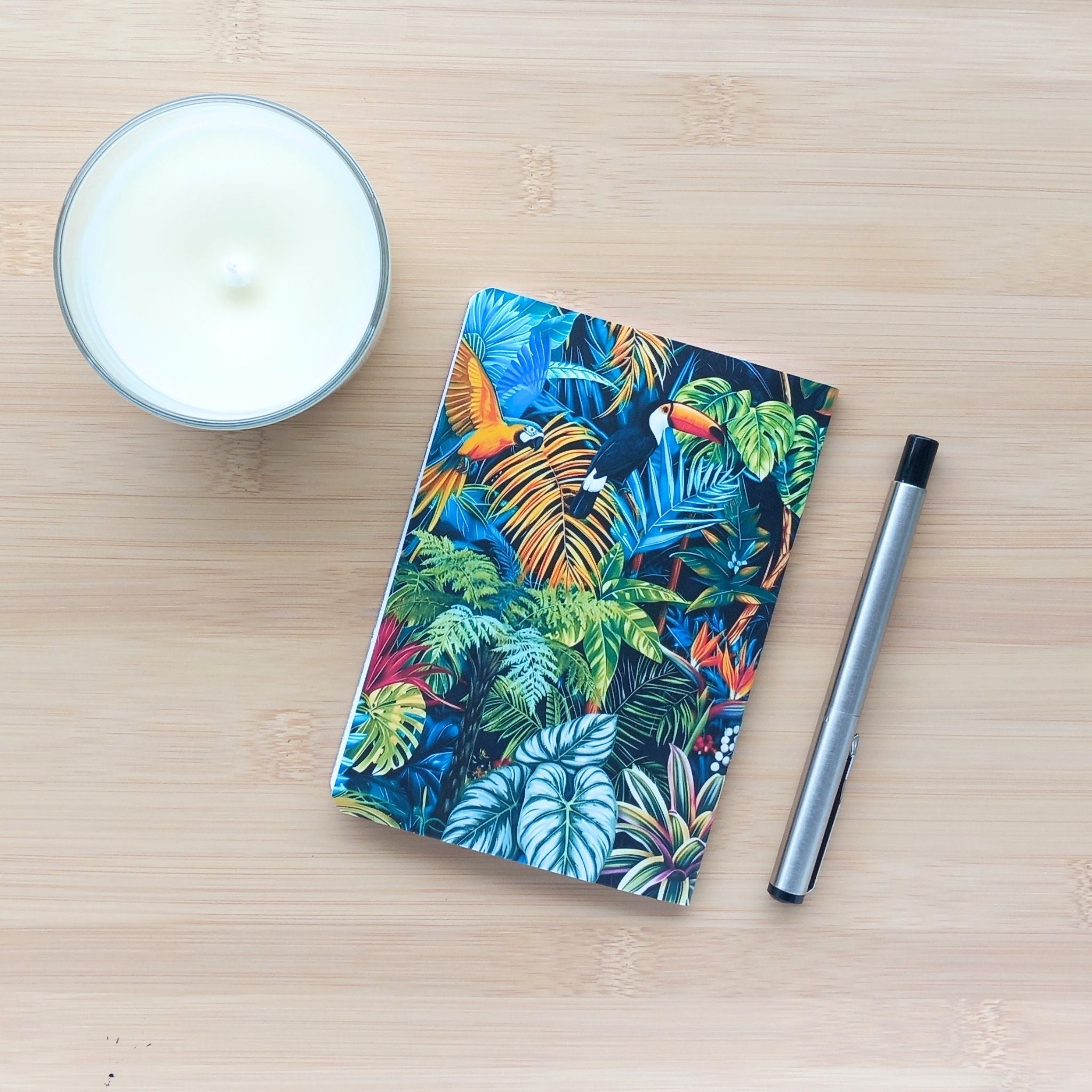 Vue de dessus de la quatrième de couverture du carnet "Maximalisme Tropical". L'illustration dense présente un feuillage exotique luxuriant abritant un toucan et un perroquet ara jaune et bleu. Le carnet de poche est posé sur un bureau en bambou à côté d'une bougie blanche et d'un stylo.