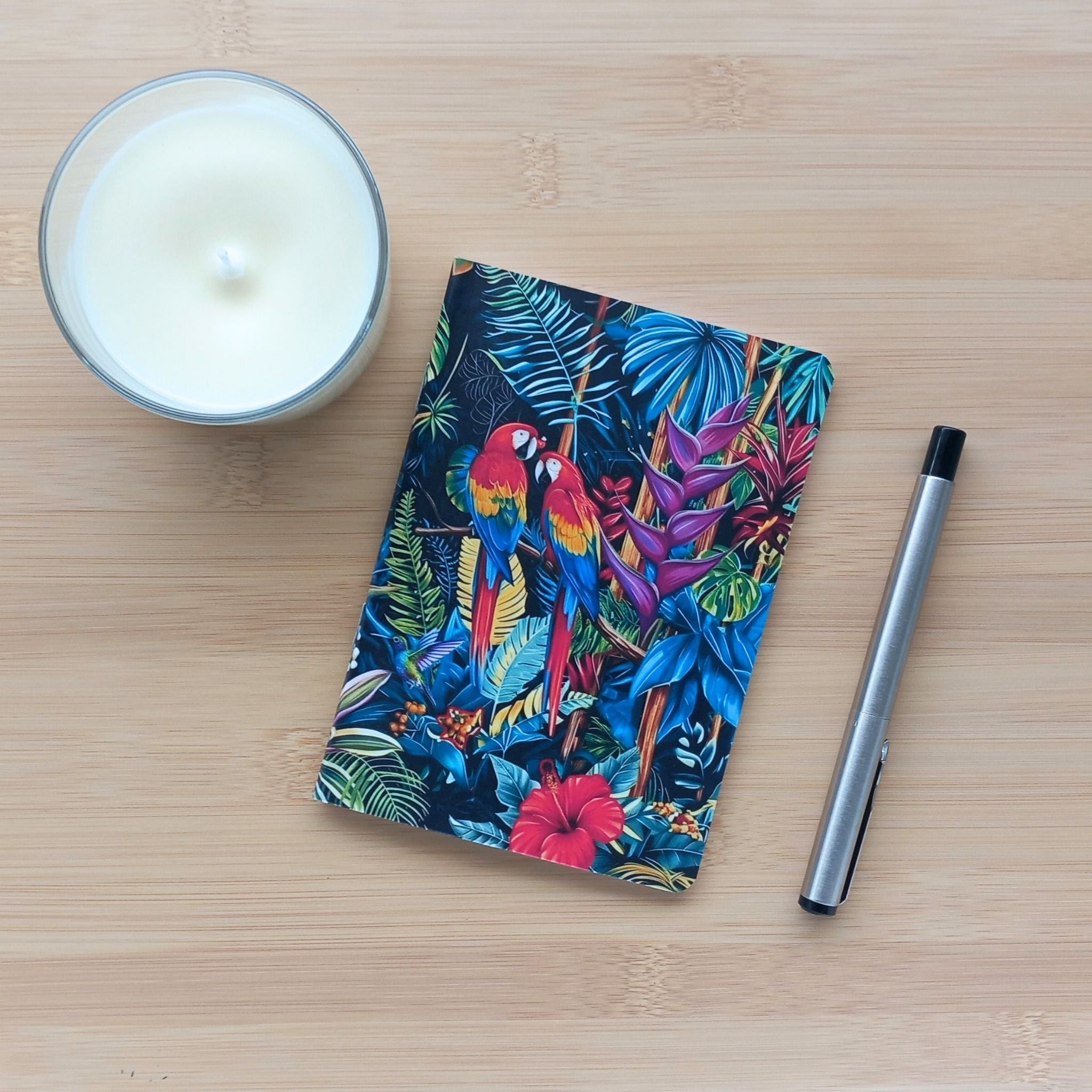 Première de couverture du carnet de notes A6 fait main, décorée d'un motif jungle foisonnant. Le design met en avant deux perroquets aras rouges perchés au milieu de grandes feuilles tropicales et d'un hibiscus. La papeterie est photographiée avec un stylo argenté et une bougie.