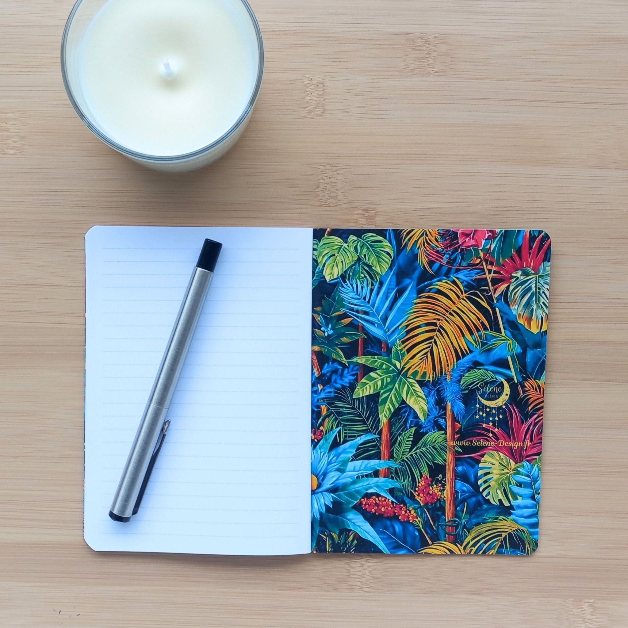 Carnet d'écriture ouvert révélant sa page de garde arrière à droite, richement illustrée de plantes tropicales vibrantes et du logo Sélène Design. Sur la page de gauche, un stylo en métal repose sur le papier ligné vierge. L'ensemble est posé près d'une bougie sur du bois clair.
