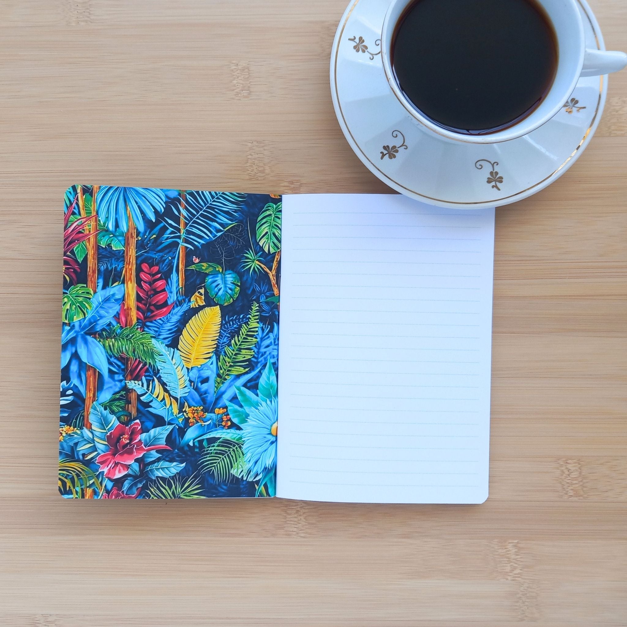 Carnet de notes tropical ouvert à côté d'une tasse de café. La couverture intérieure gauche dévoile une illustration botanique immersive aux teintes bleues, vertes et jaunes avec des fleurs exotiques. La page de droite montre le papier blanc à lignes prêt pour la prise de notes.