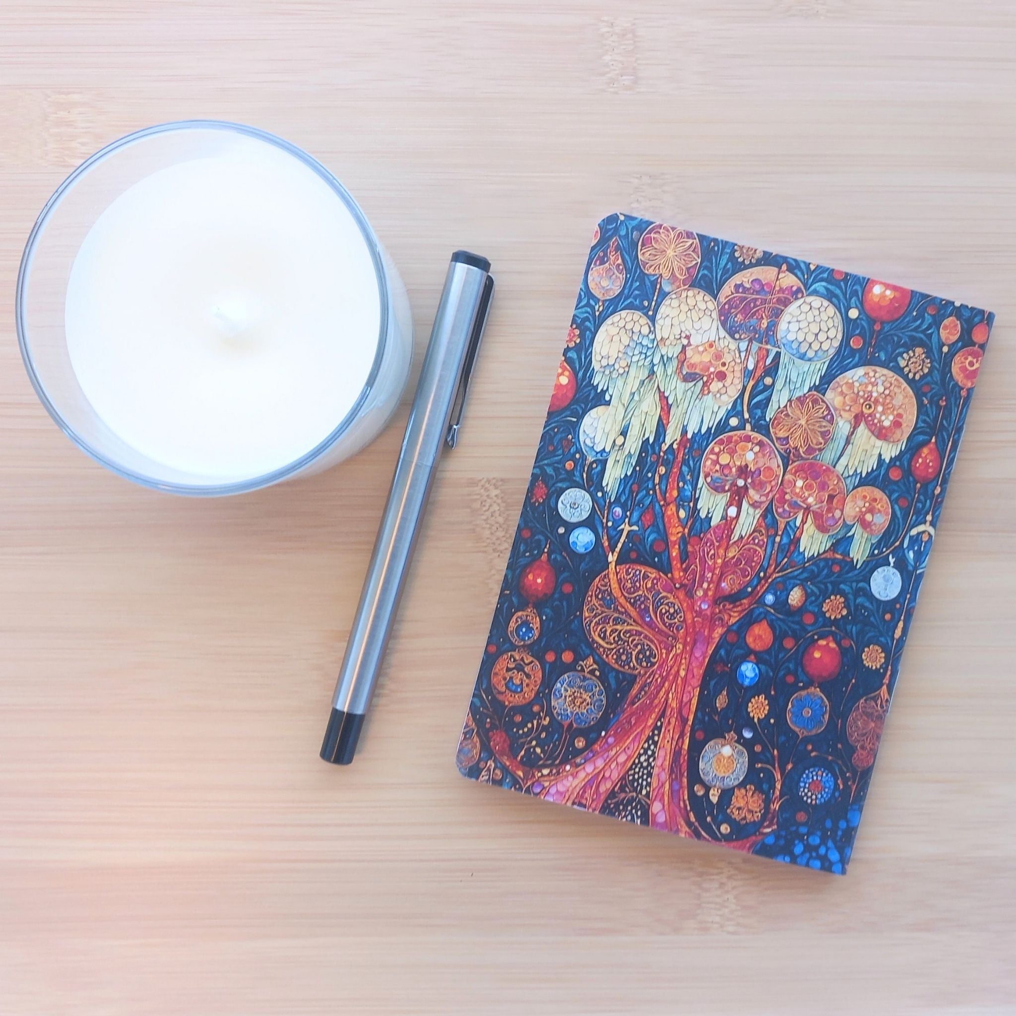 Vue de dessus de la quatrième de couverture du carnet "Songe de l'Ange". L'illustration onirique sur fond sombre présente des arbres stylisés ornés de sphères lumineuses et de motifs dorés complexes. Le carnet est posé sur une table en bois clair à côté d'une bougie et d'un stylo argenté.