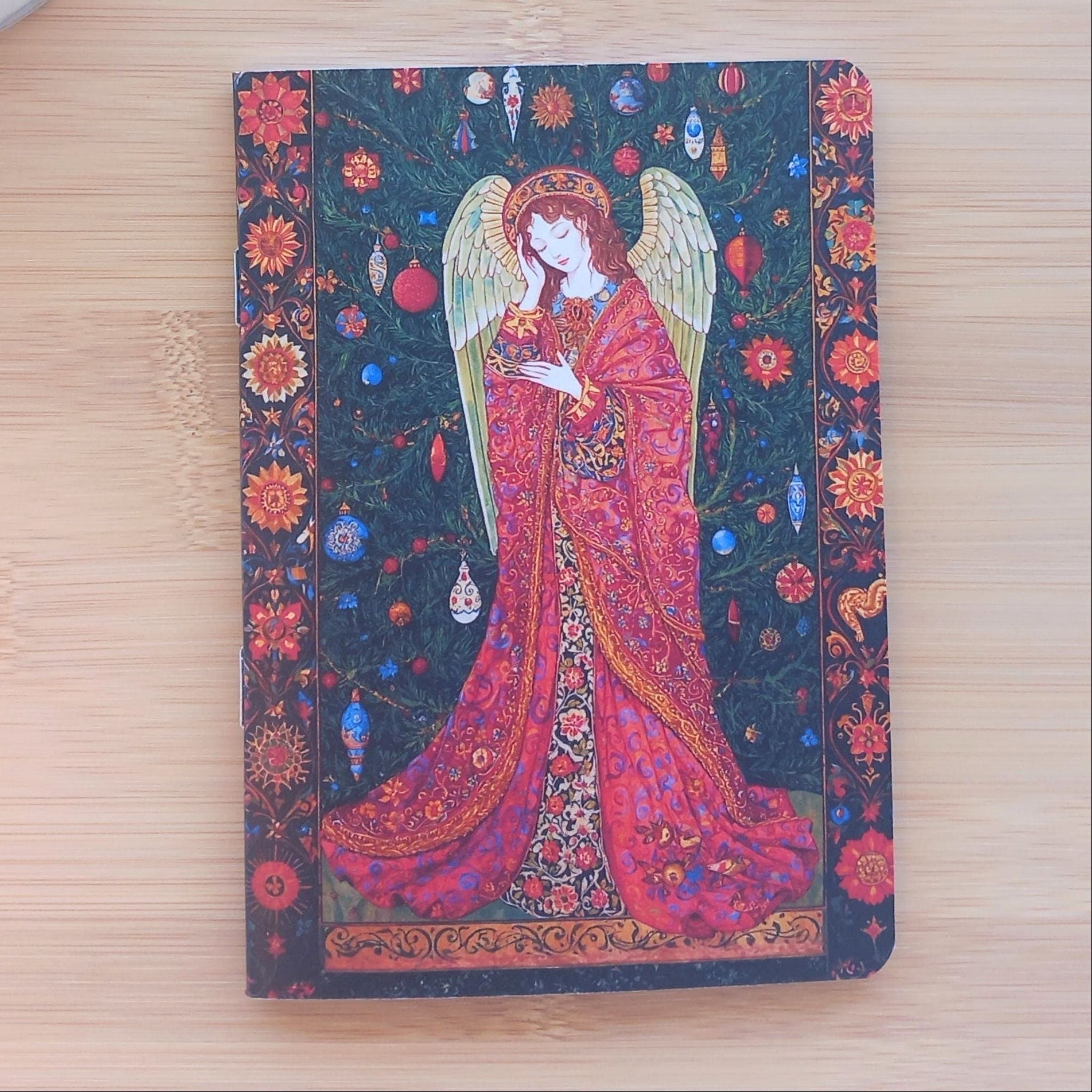 Gros plan sur le carnet de poche "Songe de l'Ange". L'illustration détaillée montre une figure angélique endormie sur un fond vert sapin parsemé de boules décoratives suspendues, évoquant la magie des fêtes et les enluminures traditionnelles.