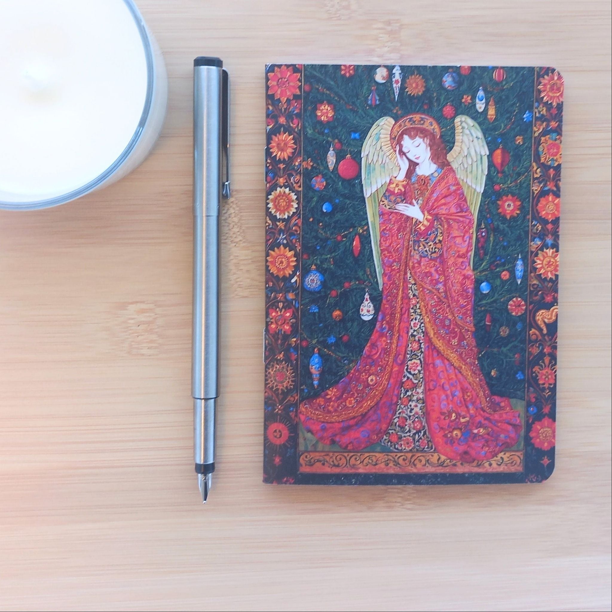 Première de couverture du carnet A6 artisanal, photographié avec un stylo et une bougie. Le design féérique met en scène un ange aux ailes blanches vêtu d'une longue robe rouge à motifs, entouré de décorations de Noël et encadré par une riche bordure florale.