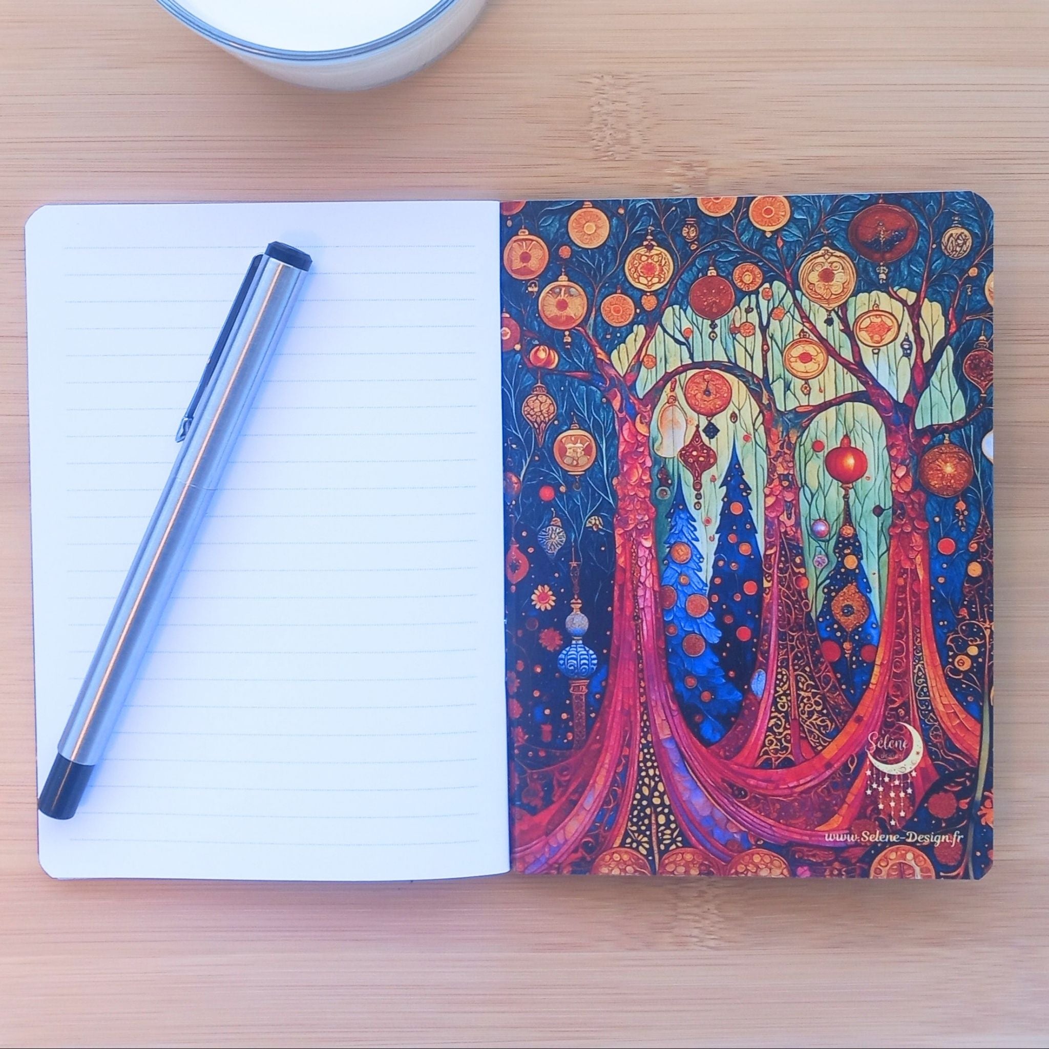 Carnet d'écriture ouvert révélant sa page de garde arrière illustrée à droite. Le dessin plonge dans une forêt enchantée aux troncs rouges et bleus, parsemée d'ornements suspendus. Sur la page lignée de gauche repose un stylo en métal, près d'une bougie.